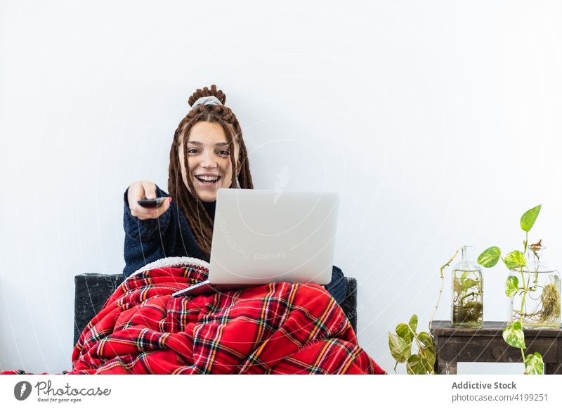 Frau mit Dreadlocks, die zu Hause an einem Laptop arbeitet und eine Fernbedienung in der Hand hat heimwärts Computer Sofa Sitzen Raum Internet