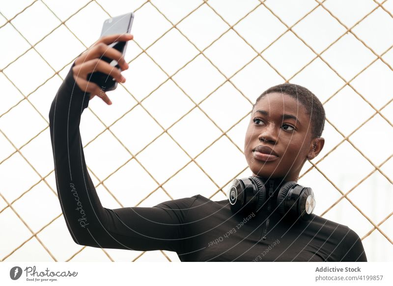 Charmante schwarze Frau mit Kopfhörern nimmt Selfie auf Smartphone Selbstportrait Gedächtnis Moment charmant Zaun Porträt benutzend Apparatur sanft feminin