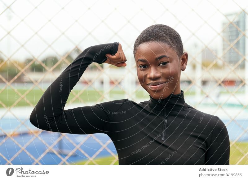 Fröhliche schwarze Frau zeigt ihre Muskeln am Gitterzaun stark Kraft Lächeln feminin sanft offen Stadt Porträt zeigen gestikulieren Sportlerin Energie Wellness