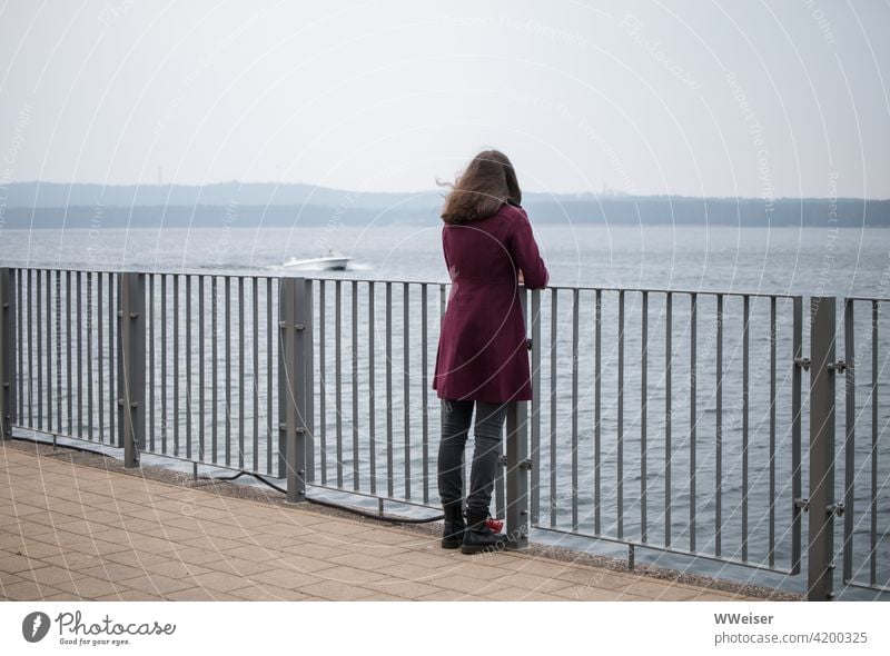 Kühler, windiger Tag. Die junge Frau schaut aufs Wasser, ein Boot brummt vorbei. Ufer Geländer Mädchen Schiff See Fluss Aussicht Diagonale Wind kühl offen