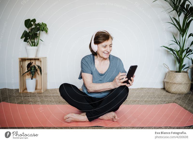 reifen kaukasischen Senior Frau übt Yoga-Pose zu Hause. mit Handy und Musik hören auf Kopfhörer. Technologie und gesunder Lebensstil Technik & Technologie
