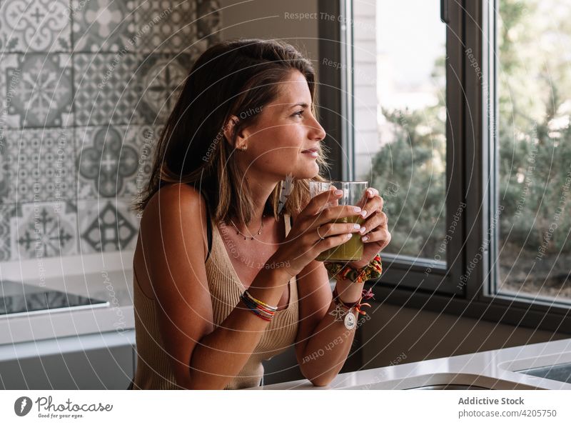 Ein kaukasisches Mädchen trinkt vor ihrem großen Küchenfenster einen grünen Gemüsesaft. Frau abgestimmt schütteln Layout Tischplatte Sellerie frisch Salat weich