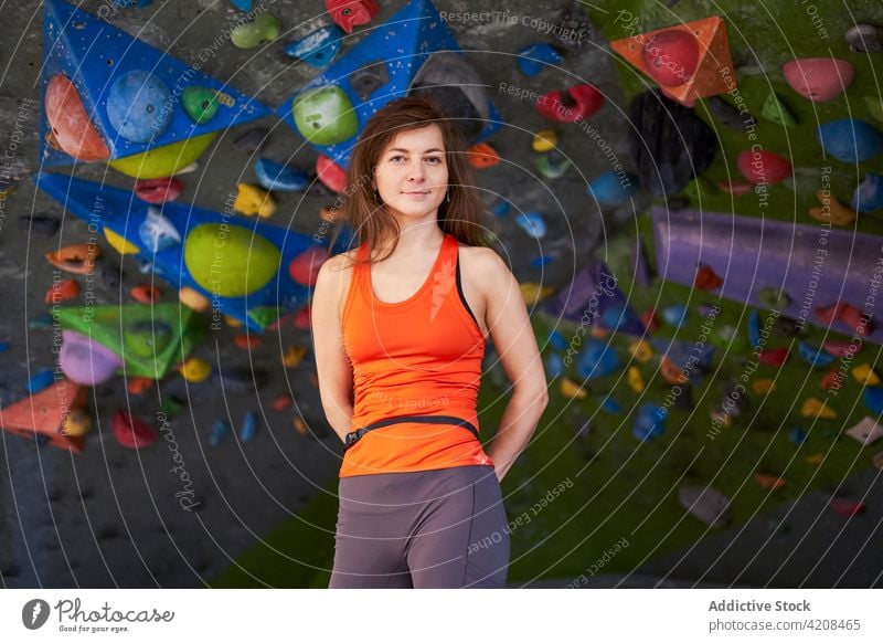 Sportlerin steht in der Nähe der Kletterwand in einer Turnhalle Hände hinter dem Rücken Bergsteiger Aufsteiger Wand Fitnessstudio Lächeln selbstbewusst passen
