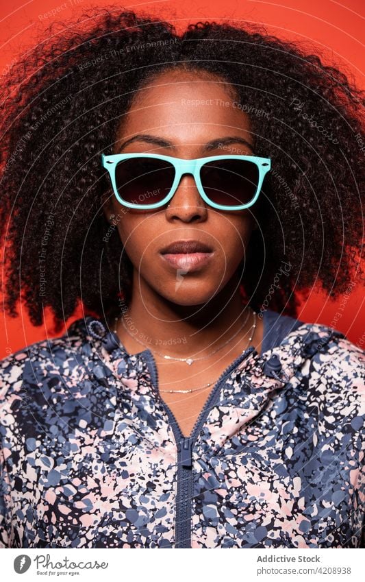 Stilvolle schwarze Frau mit Sonnenbrille vor rotem Hintergrund selbstbewusst cool Outfit Vorschein unabhängig Windstille trendy Streetstyle Accessoire ernst