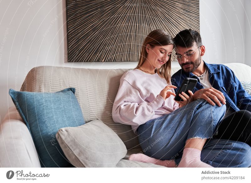 Positives junges gemischtrassiges Paar, das sich ein Smartphone auf der Couch teilt benutzend Sofa positiv romantisch Zusammensein Partnerschaft ruhen Liebe