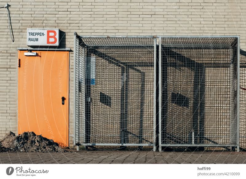 orange Tür zum Treppenhaus mit Gitterboxen auf einer Baustelle gitterbox Schattenwurf Zaun Barriere Metall Zugang Notausgang Fluchtweg Sicherheit Bauzaun