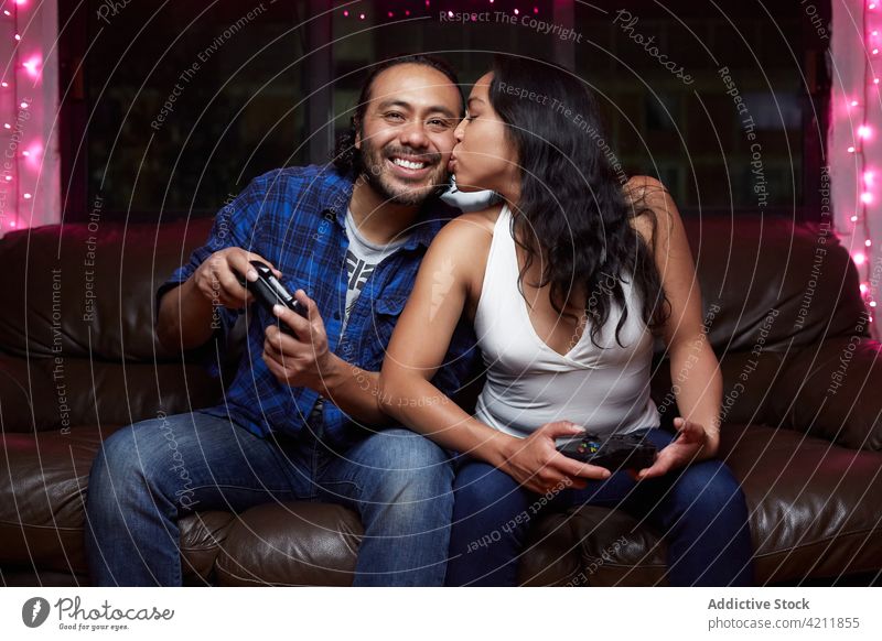 Entzücktes ethnisches Paar spielt zusammen ein Videospiel Freude spielen Kuss aufgeregt Liege Steuerkreuz Spaß haben unterhalten Zusammensein Lachen Gamepad
