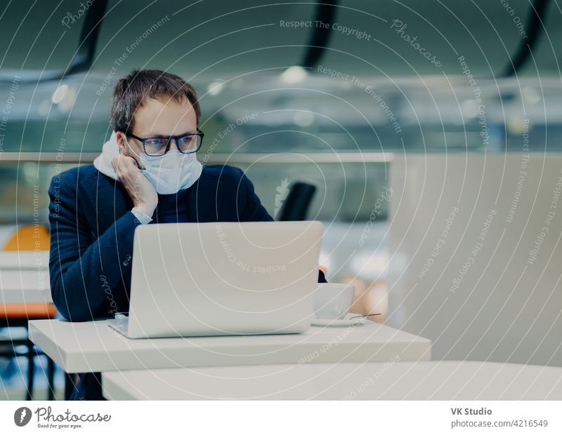 Foto eines ernsten Mannes, der sich auf dem Bildschirm eines Laptops konzentriert, arbeitet während des Ausbruchs des Coronavirus aus der Ferne, trägt eine Schutzmaske, um die Krankheit nicht zu verbreiten, trinkt Kaffee, sitzt an einem weißen Tisch