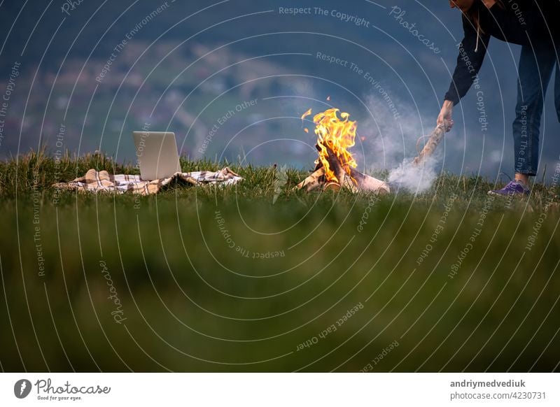 junge Frau Freiberufler wirft Brennholz in ein Feuer in den Bergen am Abend. Tourist Mädchen ruht und arbeitet im Freien Laptop Sitzen außerhalb reisen