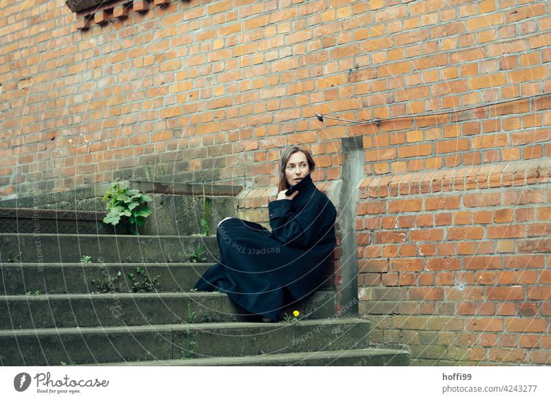 die Frau sitzt in einen Mantel gehüllt auf einer Treppe und blick über die Schulter den Weg zurück Junge Frau Melancholie Frauengesicht feminin langes Haar