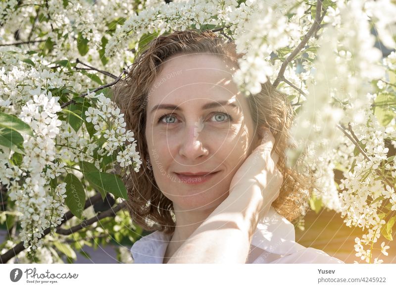 Schöne lächelnde kaukasische Frau mit lockigem Haar posiert unter einem blühenden Baum. Frühling Fotoshooting, sonniger Tag. Porträt einer glücklichen Frau. Frühlingssonne