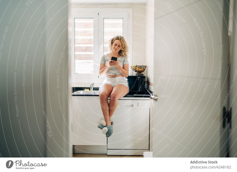Fröhliche Frau mit Smartphone auf dem Küchentisch sitzend benutzend Browsen Inhalt Apparatur Glück Telefon modern Lächeln jung Gerät positiv Stil lässig Moment