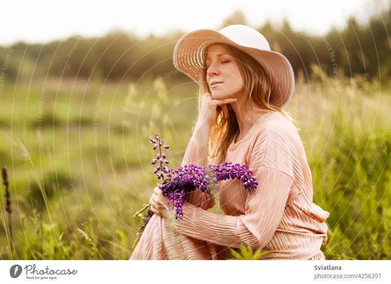 Porträt einer schönen Blondine mit einem Strauß Lupinen in den Händen auf dem Rasen sitzend Mädchen Feld Blumenstrauß Hut Sommer ein Blumenstrauß aus Lupinen