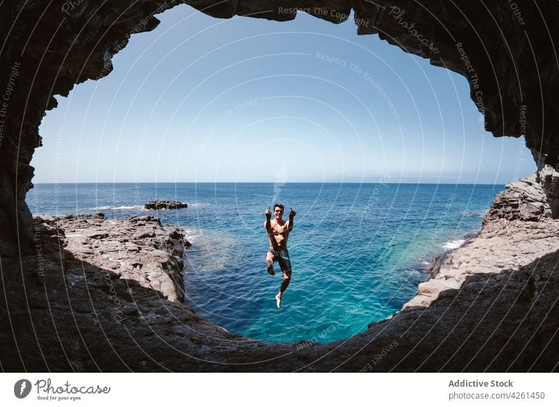Lächelnder Mann zeigt Fickgesten beim Sprung über das Meer springen Höhle ficken Inhalt Natur Hochland Golfloch Himmel Aktivität Energie zeigen gestikulieren