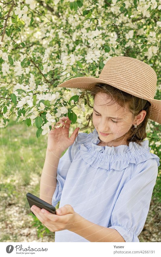 Cute schöne lächelnde Mädchen in einem Strohhut schießt Inhalte für soziale Netzwerke auf einem Smartphone. Frühling Fotorahmen vor dem Hintergrund der blühenden Apfelbäume. Glückliches Kind. Vertikale Aufnahme
