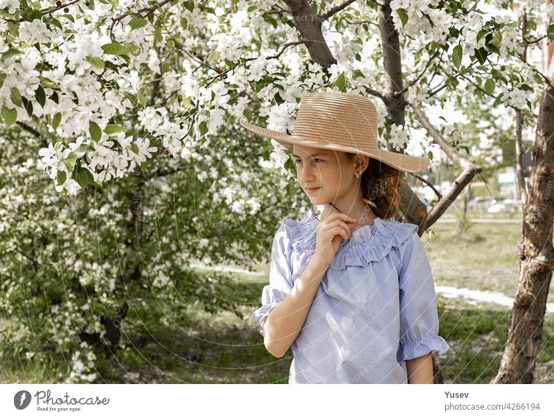 Porträt eines niedlichen, nachdenklichen Mädchens mit Strohhut in einem blühenden Garten. Frühling Fotoaufnahme vor dem Hintergrund der blühenden Apfelbäume. Glückliches Kind. Platz kopieren