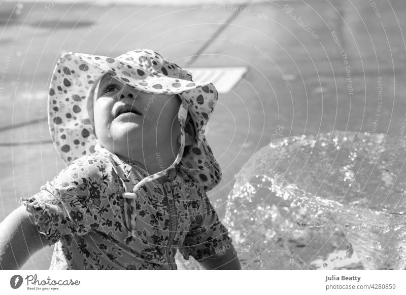 Ein Jahr altes Baby in einem Wasserpark an einem heißen Tag; Kleinkind trägt Sonnenschutz UPF Sonnenhut und Badeanzug, wie sie schaut zu den Eltern für Beruhigung