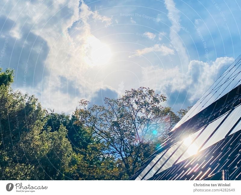 Die Sonne scheint hell vom leicht bewölkten Himmel und spiegelt sich in den Kollektoren der Solaranlage und der Photovoltaikanlage eines Wohnhauses Solarthermie