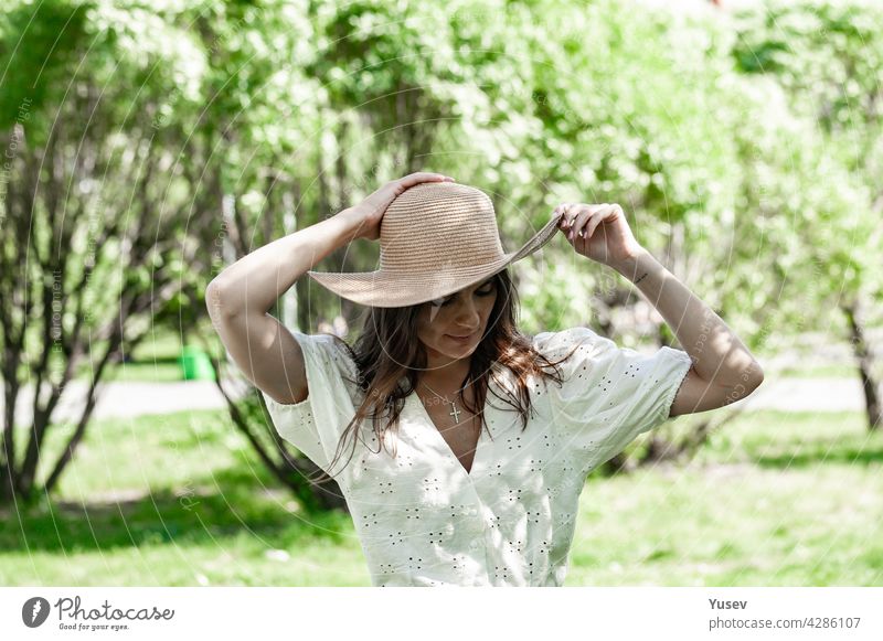 Junge schöne glückliche Frau in modischen Strohhut. Eine brünette Frau in einem weißen Hemd stehen im Freien und hält ihren Hut mit den Händen. Blühende Frühling Apfelbaum Park Hintergrund. Sonniger Sommertag