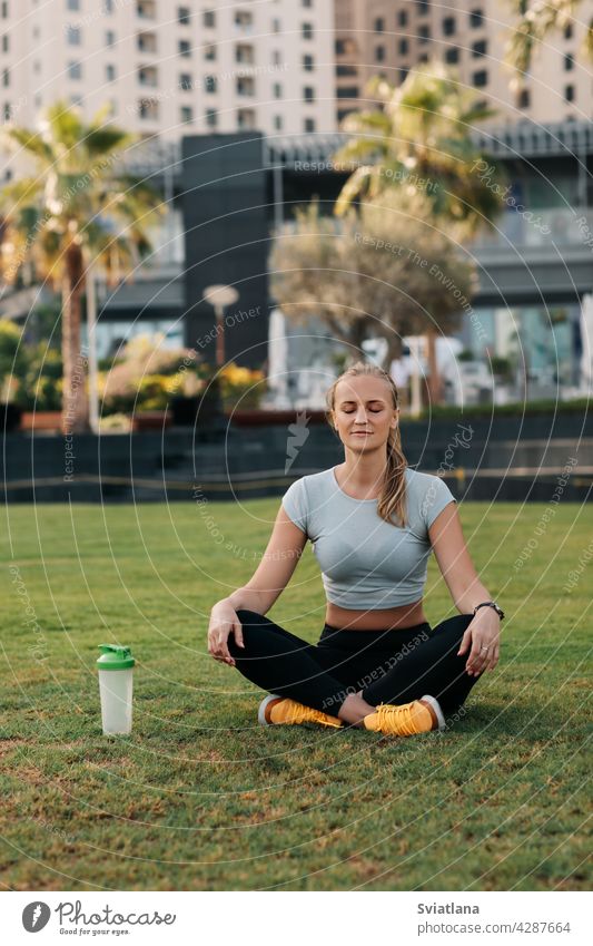 Eine junge Frau sitzt im Lotussitz auf dem Rasen im Park neben dem Haus und meditiert nach einem Workout. Sport, Fitness, Yoga. Gesunder Lebensstil Konzept