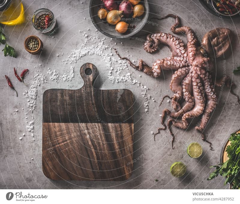 Mediterranes Lebensmittelkonzept mit rohem Tintenfisch, Gewürzen, Gemüse und Kräutern. Kochvorbereitung mit frischen Zutaten und Geräten. Rustikales Schneidebrett auf grauem Betonhintergrund. Ansicht von oben