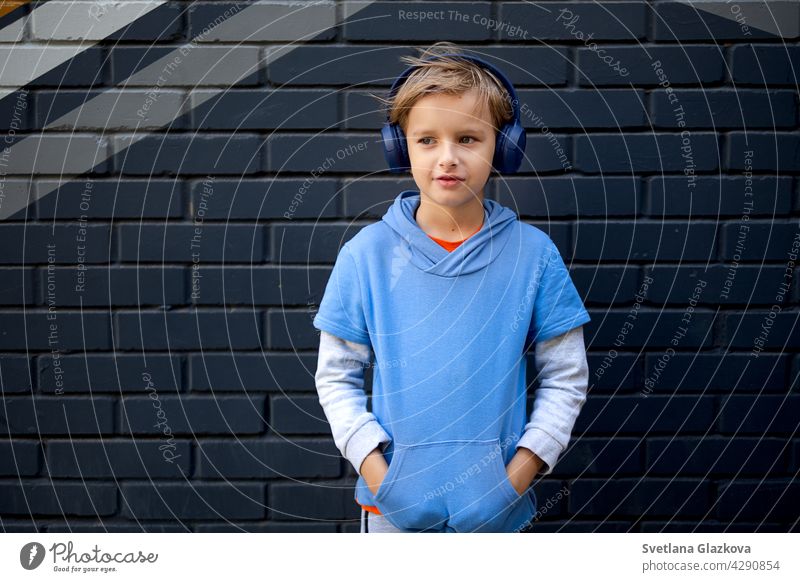 Blonde kaukasische Junge in einem blauen Kapuzenpulli und Kopfhörer steht auf einer Straße in der Stadt gegen eine graue Backsteinmauer, Musik hören in einer guten Stimmung
