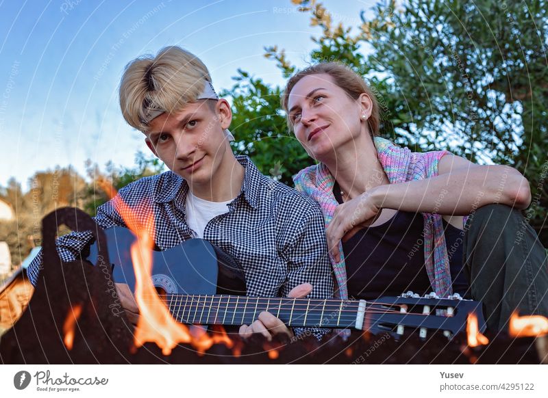 Glückliche fröhliche Mutter und Sohn sitzen im Hinterhof in der Nähe des Feuers und spielen eine akustische Gitarre. Sie singen Lieder und genießen einen Sommerabend. Sommer-Lifestyle-Fotografie. Blauer Himmel Hintergrund.