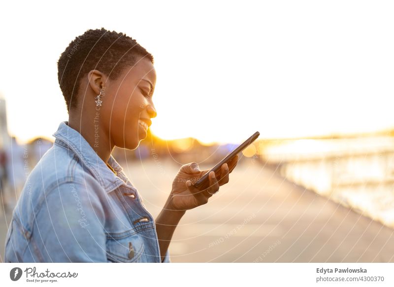 Lächelnde junge Frau mit Smartphone im Freien Afro-Look stolz echte Menschen Stadtleben Afroamerikaner Afrikanisch Schüler schwarz Ethnizität sonnig