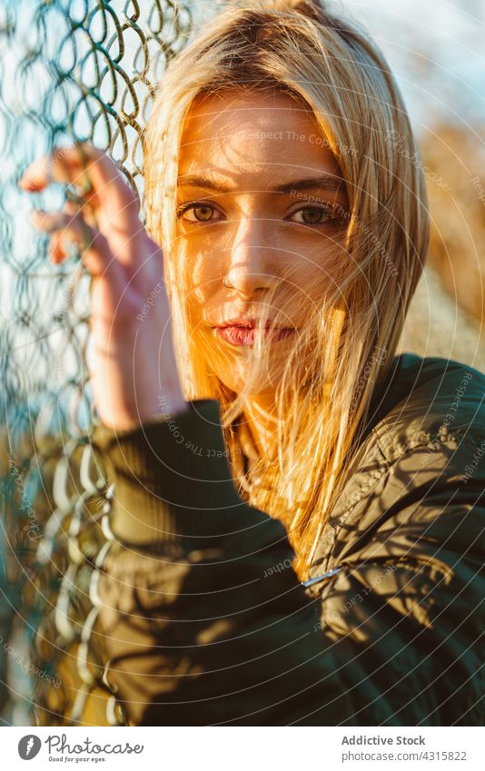 Ruhige Frau lehnt sich im Sonnenlicht an einen Netzzaun Zaun Porträt Kettenglied Windstille blond charmant modern Schönheit jung hell sorgenfrei allein Jacke