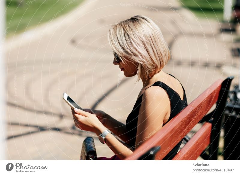 Eine junge Frau macht ein Selfie auf einer Parkbank sitzend, passt ihr Make-up und ihre Frisur an und betrachtet dabei ihr Spiegelbild auf dem Bildschirm Bank