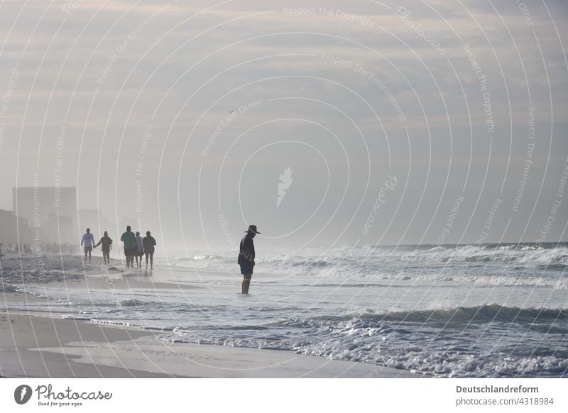 Mann mit Hut betrachtet Wellen am Strand. Mehrere Menschen spazieren im Hintergrund. Person kurze Hose Jacke Sand Morgens frueh Sonnenaufgang dunst Küste Meer