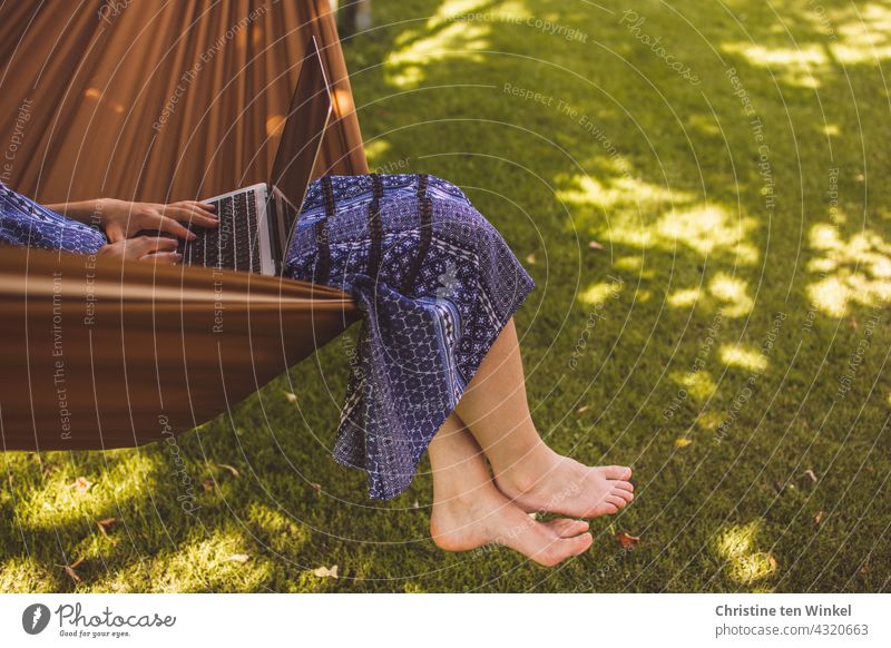 Eine junge Frau im blauen Sommerkleid sitzt gemütlich in einer Hängematte im Garten, arbeitet am Notebook und lässt die Füße baumeln laptop Computer Homeoffice