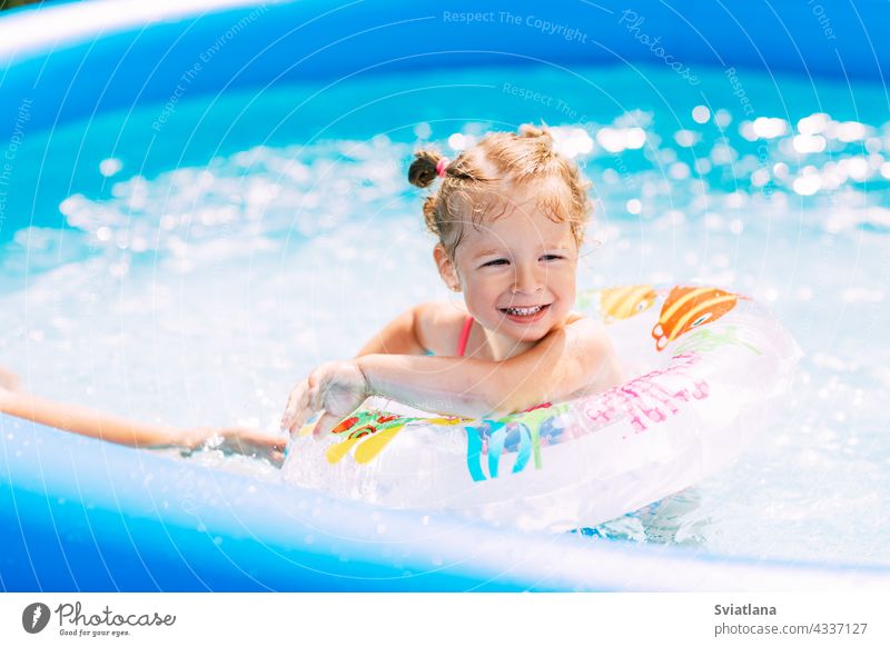 Ein lachendes Baby im hellen Badeanzug lernt mit Hilfe eines aufblasbaren Kreises das Schwimmen im Pool im Garten. Sommerzeit, Erholung, Rückansicht Mädchen
