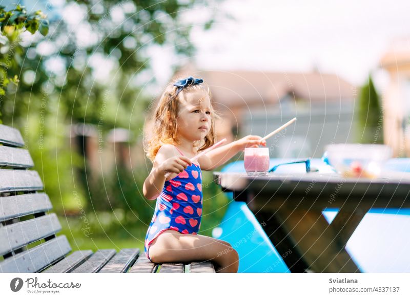 Ein süßes Baby trinkt einen Beeren-Smoothie durch einen Strohhalm und sitzt auf einer Bank im Garten. Erholung im Freien, Sommerferien für Kinder Gesundheit