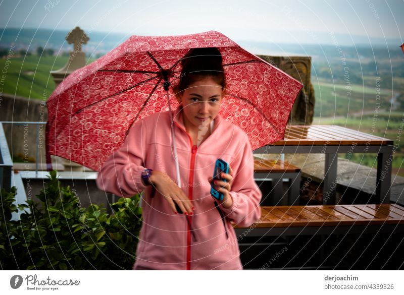 Alles in Rosa. Junges Girl steht mit Schirm und Regenjacke im Außenbereich des  Restaurant.  Das Handy in der Hand und schaut verzweifelt zu dem Fotografen,  wann ist  er endlich fertig .