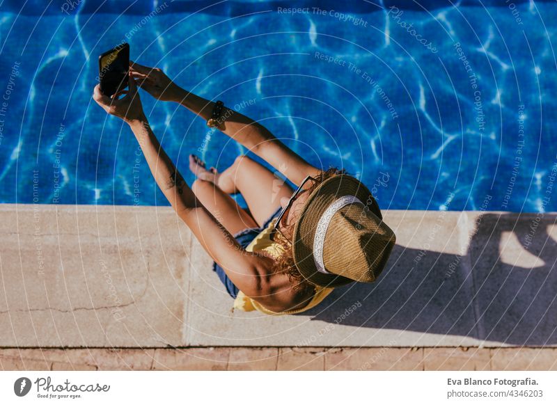 Draufsicht auf junge kaukasische Frau, die am Pool sitzt und ihr Handy benutzt, um ein Selfie zu machen. Sommerzeit, Urlaub und Lebensstil Technik & Technologie