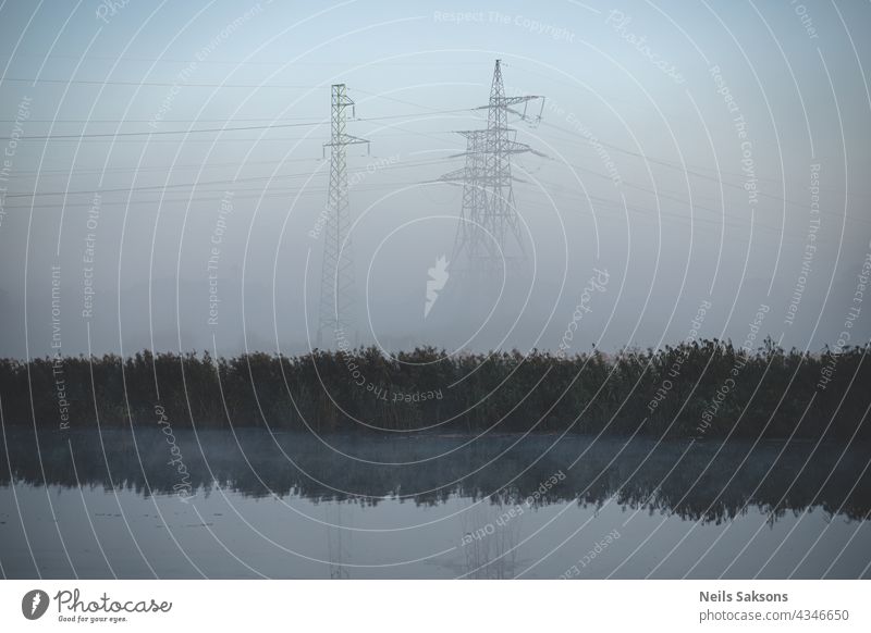 elektrische Transitlinien und Pylonen in der Nähe von Fluss in nebligen Morgen in Lettland. Viele trockene Schilf im Oktober reflektiert im Wasser. Herbst kalten Morgennebel im Hintergrund.