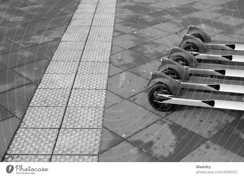 Moderne E-Roller aufgereiht zur Vermietung neben dem taktilen Leitsystem für Sehbehinderte auf dem Straßenpflaster in der Innenstadt von Frankfurt am Main in Hessen, fotografiert in neorealistischem Schwarzweiß