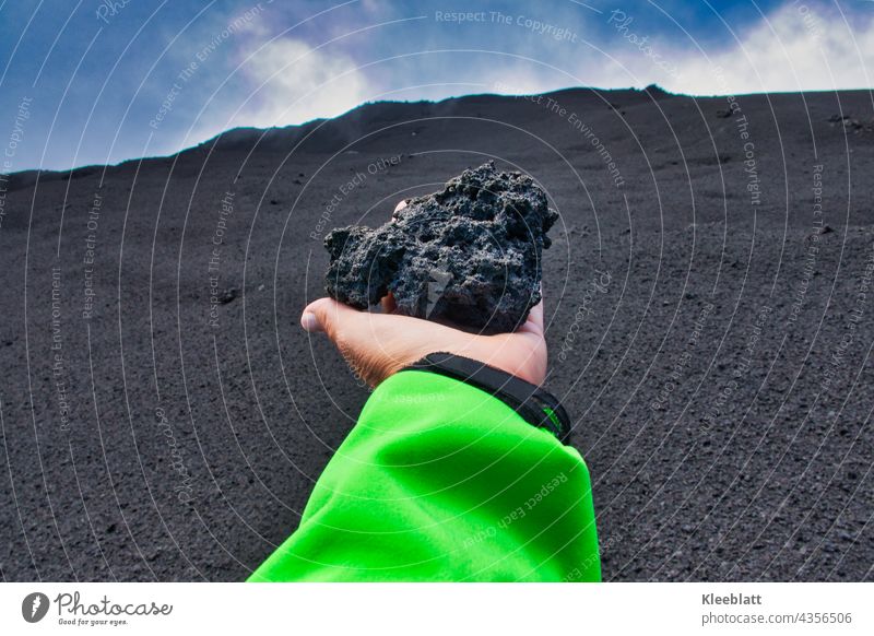 Ätna - Frauenhand hält einen großen Vulkanaschebrocken in der ausgestreckten Hand  Richtung Vulkankrater Grüne Jacke blauer Himmer Weiße Wolken schwarze Asche,