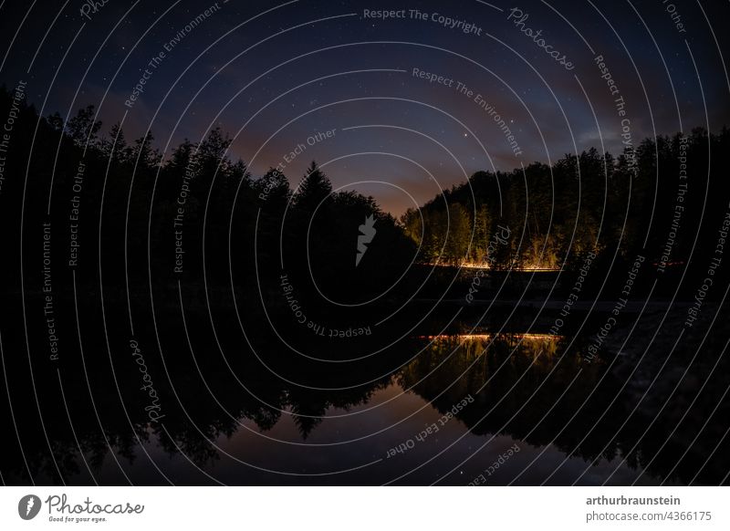 Nachtaufnahme von ruhigem Wasser unter Sternenhimmel mit Autolichtern in der Ferne im Wald natur Außenaufnahme Menschenleer Landschaft nacht see wasser sterne