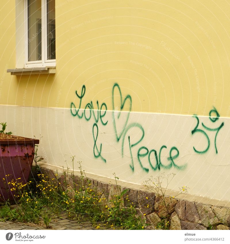 Love & Peace steht in grüner Schrift an einer gelben Hauswand neben einem abgestellten Container / Graffito love and peace Liebe Frieden Graffiti Schmiererei