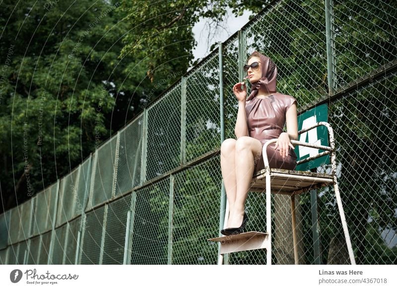 Neben einem rostigen Zaun eines Tennisspielplatzes sitzt ein wunderschönes Model. Sie trägt ein rosa Latexkleid, eine modische Sonnenbrille und natürlich hohe Absätze. Meine Art von Richterin, die die ganze schöne Arbeit macht.