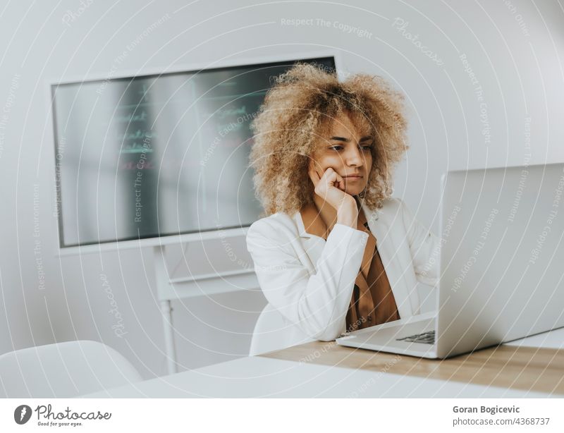 Junge Frau mit lockigem Haar arbeitet an einem Laptop in einem hellen Büro mit großem Bildschirm hinter ihr Erwachsener attraktiv schön hinten schwarz Business