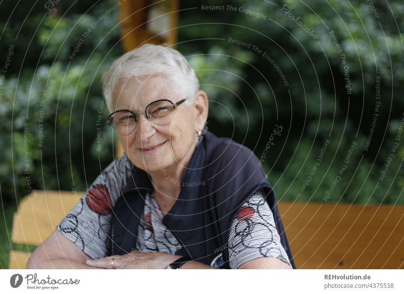 Seniorin sitzt am Waldrand und lächelt 60 und älter Porträt Weiblicher Senior Frau Großmutter weißhaarig grauhaarig natürlich authentisch Brille Natur alt