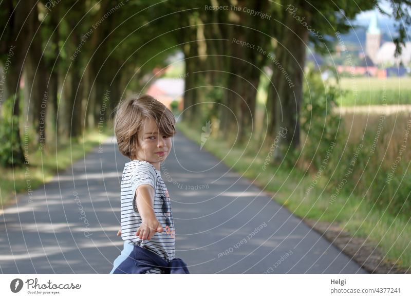 Junge steht auf einer Alleestraße, blickt und zeigt zum Fotografen Mensch Kind Kindheit Straße Baum Spaziergang draußen unterwegs Außenaufnahme Wege & Pfade