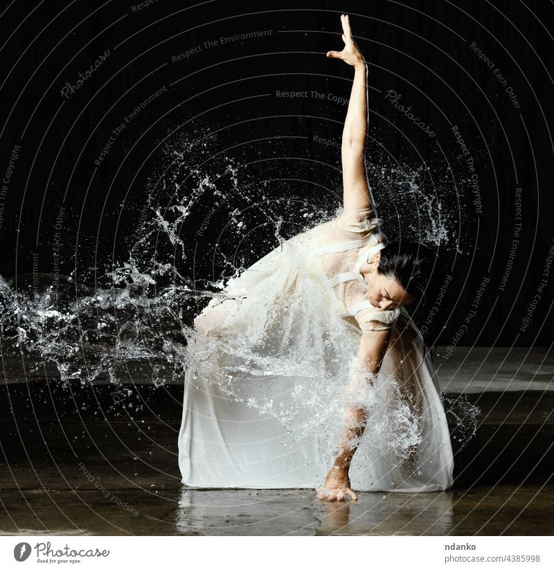 schöne Frau kaukasischen Aussehens mit schwarzen Haaren tanzt in Wassertropfen auf schwarzem Hintergrund. Die Frau trägt ein weißes Chiffonkleid beweglich aqua