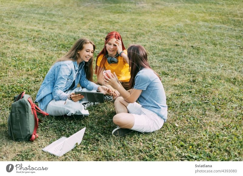 Teenager-Mädchen sitzen mit einem Rucksack und einem digitalen Tablet auf einer grünen Wiese in einem Park und bereiten ein gemeinsames Projekt vor. Bildung und Wissen, Lebensstil von Teenagern