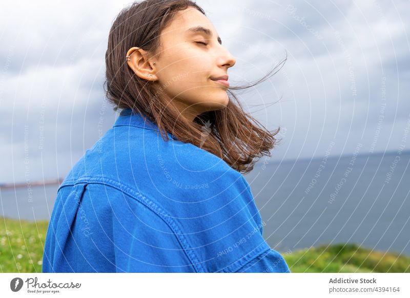 Zufriedene Frau auf einem Hügel am Meer sitzend Augen geschlossen MEER genießen Seeküste wolkig bewundern Lächeln Strandpromenade Harmonie Natur Himmel grün