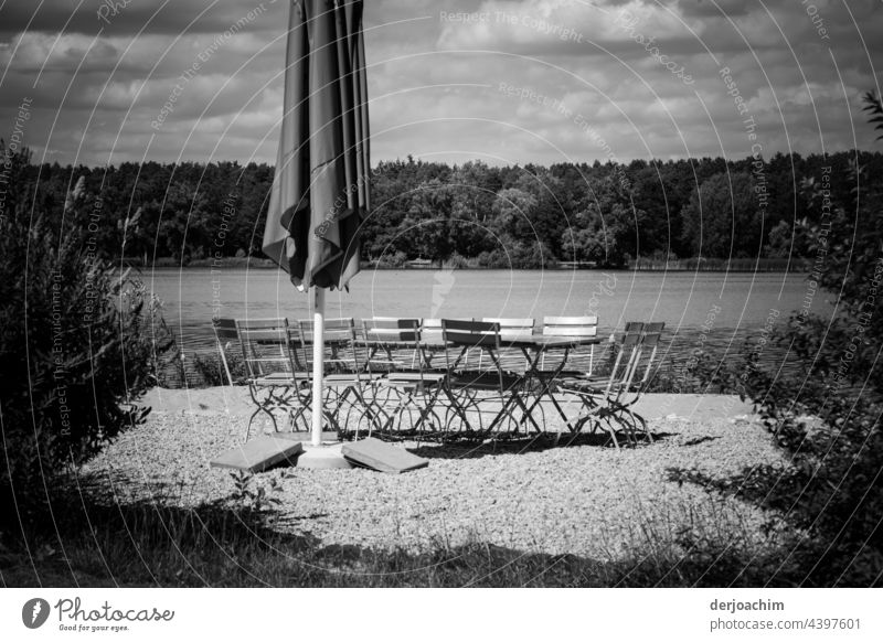 Ein eingeklappter Sonnenschirm,  ein paar leere Stühle und Tisch , sowie das Wasser von einem See und im Hintergrund ganz viel Wald. Zu allem am Himmel viele Wolken. Jetzt fehlen nur noch die Gäste...
