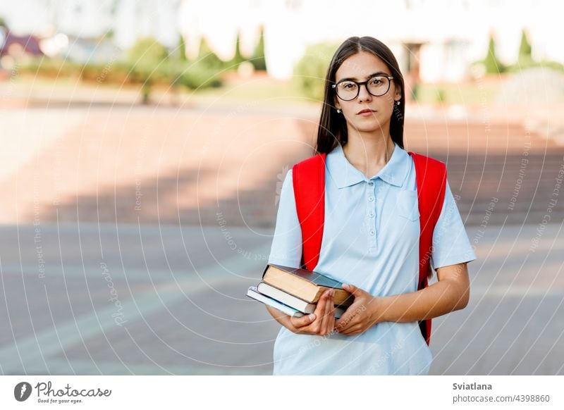 Eine lächelnde Studentin mit Brille, Büchern und einem roten Rucksack. Porträt einer Studentin vor einem städtischen Hintergrund. Das Konzept der Bildung